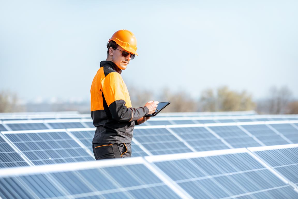 Engenheiro em roupas de trabalho de proteção realizando manutenção de painéis solares com tablet digital em uma planta fotovoltaica no telhado. Conceito de manutenção e configuração de usina de energia solar